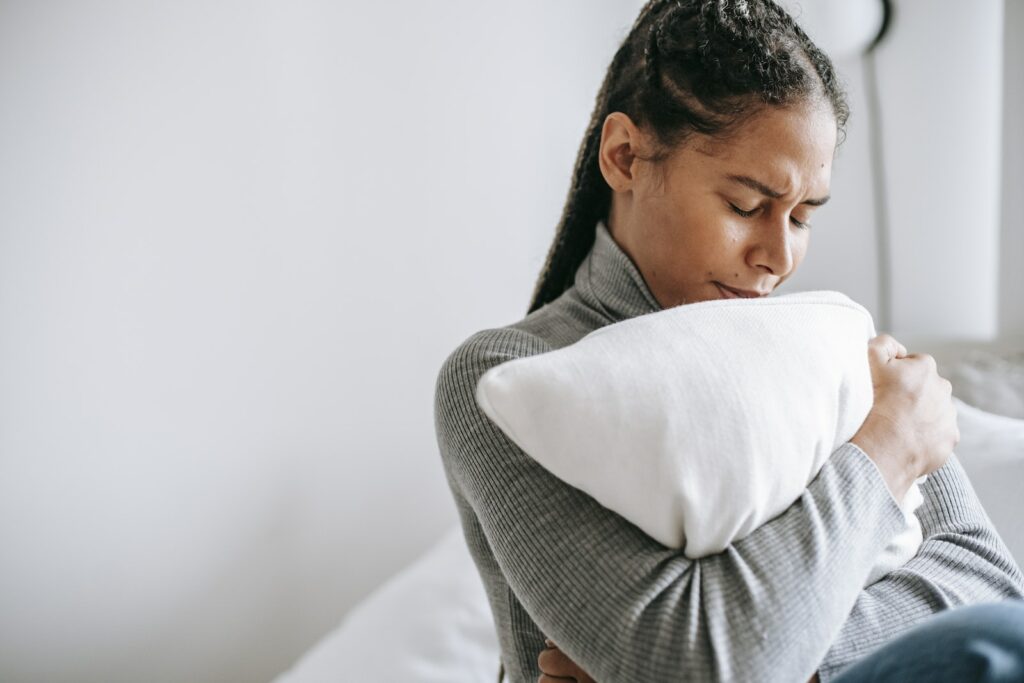 Young ethnic female with closed eyes and sad face expression sitting on bed and embracing pillow